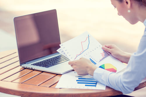 Woman sitting at table, with laptop and looking at charts on paper.